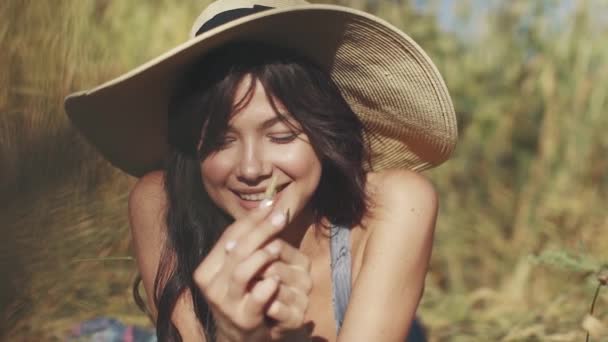 Close-up πορτρέτο του ένα κορίτσι του χωριού σε ένα ψάθινο καπέλο με ένα Καλαμάκι στα χέρια της. Ένα χαριτωμένο κορίτσι που χαμογελά και φαίνεται στην κάμερα — Αρχείο Βίντεο