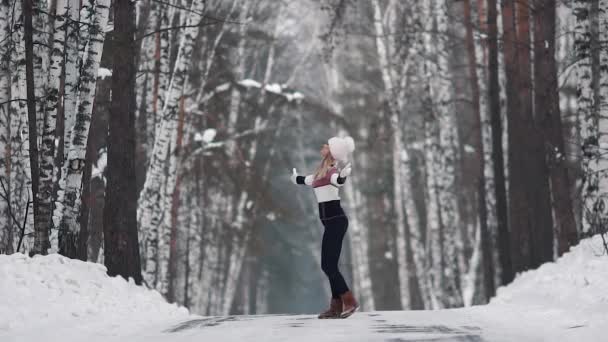 Young smiling girl is happily spinning in the winter forest. Portrait of a young woman in a knitted sweater and a mitten cap. slow motion — Stock Video