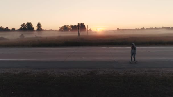 Aérea. una joven está montando en un monopatín a lo largo de una carretera desierta al amanecer — Vídeo de stock