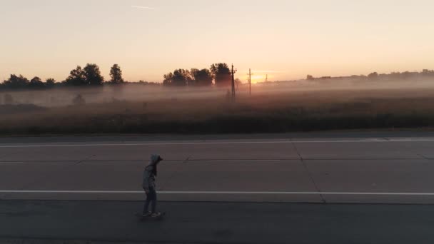Aéreo. uma adolescente patina em um skate ao longo de uma estrada deserta fora da cidade ao amanhecer. Jovem de camisola com capuz e jeans — Vídeo de Stock
