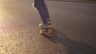 bacaklar kız kaykay yakın çekim. Bir kız spor ayakkabı ve yırtık kot günbatımında ıssız bir yolu boyunca bir kaykay sürme