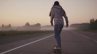 Kız paten kaykay sabah sis çerçevede bir ıssız bir otoyol üzerinde. Arkadan görünüm. ağır çekim.