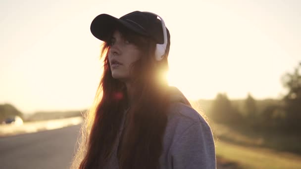 Portret ragazza dai capelli rossi ai raggi del sole nascente. capelli svolazzanti nel vento. adolescente ascolta musica con le cuffie — Video Stock