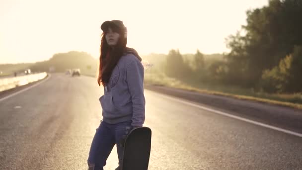 Menina está andando com um skate em suas mãos ao pôr do sol. Menina ruiva com fones de ouvido e jeans rasgados. câmara lenta — Vídeo de Stock