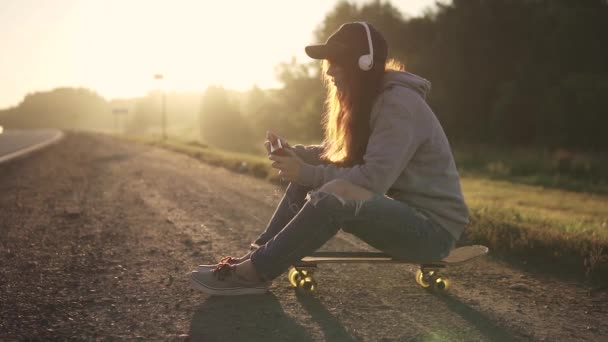 Chica pelirroja se sienta en un monopatín al lado de la carretera y escucha música en los auriculares. Una chica disfruta de la soledad al atardecer . — Vídeos de Stock