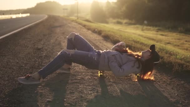Meisje hipster ligt op een skateboard langs de kant van de weg en luister naar retro muziekspeler. Portret van een roodharige meisje bij zonsondergang — Stockvideo