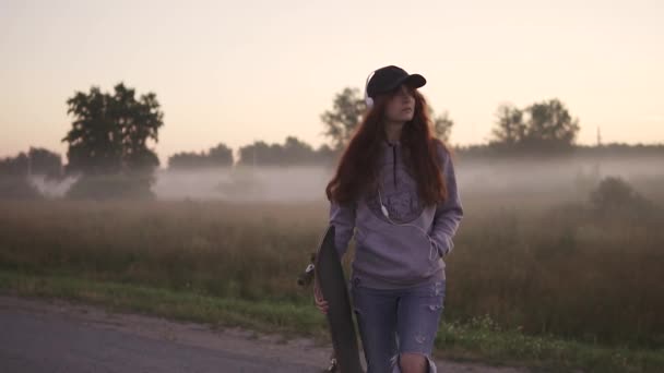 Red-haired girl with a skateboard in her hands is walking against the background of the morning mist. A hipster girl in ragged jeans and a sweatshirt with a hood listening to music on headphones — Stock Video