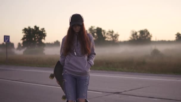 Ritratto di un primo piano hipster. La ragazza dai capelli rossi cammina lungo un'autostrada deserta con uno skateboard in mano sullo sfondo della nebbia mattutina. rallentatore . — Video Stock