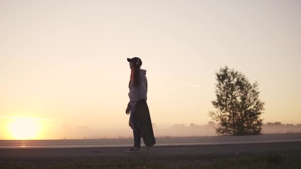 Silueta de una chica hipster con un monopatín en las manos contra el telón de fondo del sol poniente. Una chica con auriculares camina por una carretera desierta fuera de la ciudad. cámara lenta . — Vídeos de Stock