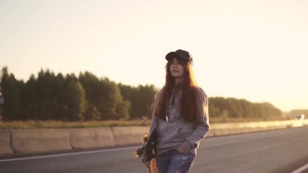 Portret van een roodharige meisje in hoofdtelefoons met een skateboard in handen wandelen langs de weg langs een verlaten snelweg bij zonsondergang. — Stockvideo