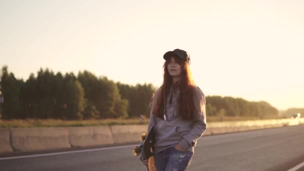 Retrato de uma menina de cabelos vermelhos em fones de ouvido com um skate nas mãos Caminhando ao longo da beira da estrada ao longo de uma estrada deserta ao pôr do sol . — Vídeo de Stock