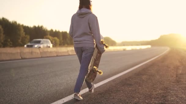 Девушка в наушниках и со скейтбордом в руках, идущей вдоль пустынного шоссе на закате. вид сзади. замедленное движение . — стоковое видео