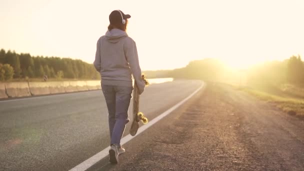 Een meisje in de hoofdtelefoon en met een skateboard in haar handen lopen langs de kant van een verlaten weg bij zonsondergang. Achteraanzicht. Slow motion. — Stockvideo