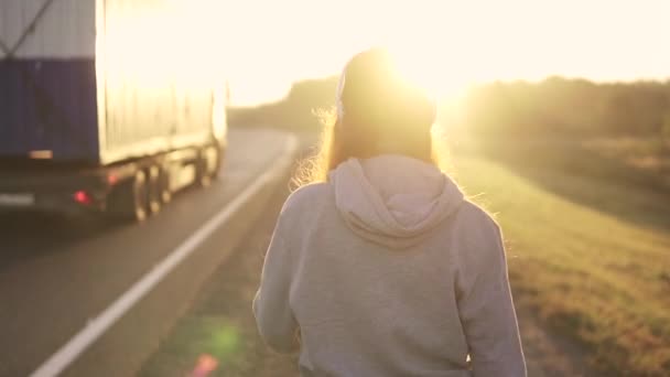 Close-up πορτρέτο της μια κοκκινομάλλα κοπέλα ακουστικά περπατώντας στην άκρη του δρόμου το ηλιοβασίλεμα. πίσω όψη. αργή κίνηση. — Αρχείο Βίντεο