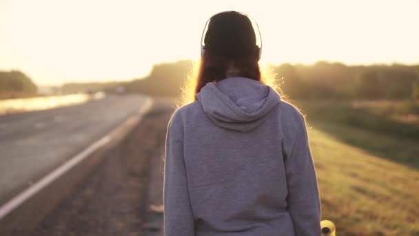 Nahaufnahme eines rothaarigen Mädchens mit Kopfhörern am Straßenrand bei Sonnenuntergang. Rückseite. Zeitlupe. — Stockvideo