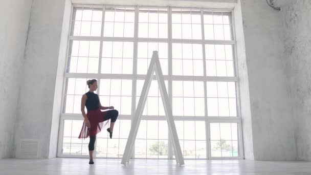 Ballerina in schwarzem Body und rotem Rock dehnt sich im Atelier vor dem Hintergrund eines großen hellen Fensters. Zeitlupe — Stockvideo