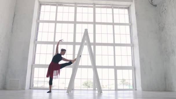 Ballerina i svart bodysuit och röd kjol gör en sträcka i studion på bakgrunden av ett stort ljust fönster. slowmotion — Stockvideo