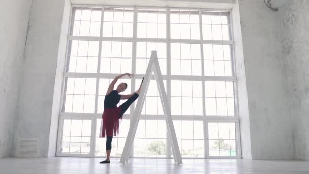 Μπαλαρίνα σε μαύρο κορμάκι με κόκκινη φούστα και κάνει ένα τέντωμα στο στούντιο για το ιστορικό της ένα μεγάλο παράθυρο φωτός. αργή κίνηση — Αρχείο Βίντεο