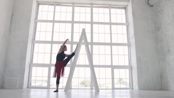 Ballerina i svart bodysuit och röd kjol gör en sträcka i studion på bakgrunden av ett stort ljust fönster. slowmotion — Stockvideo