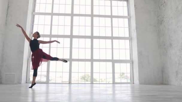 Uma bailarina em um bodysuit preto e uma saia vermelha dança um balé clássico no Estúdio contra uma grande janela de luz. câmara lenta — Vídeo de Stock