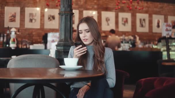 Een mooi meisje zit in een koffiehuis en texting een bericht op haar mobiele telefoon. — Stockvideo