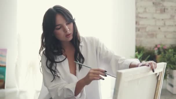 Aantrekkelijk meisje schilder schetst een beeld in een home studio. kunstenaar met een penseel in de hand — Stockvideo