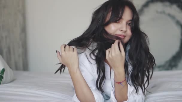 Ritratto di una bella ragazza con una camicia sdraiata sul letto. ragazza tocca i capelli e guarda misteriosamente nella fotocamera — Video Stock