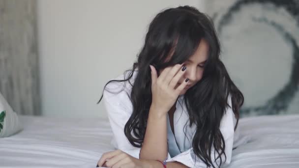 Porträt eines attraktiven Mädchens im Hemd, das auf dem Bett liegt. Mädchen berührt ihr Haar und blickt geheimnisvoll in die Kamera — Stockvideo
