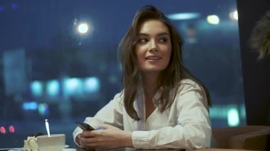 kız bir kafede tek başına otururken bir akıllı telefon kullanır. gece şehrin ışıkları arka plan üzerinde bir kafede bir genç portresi