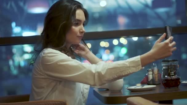 Молода дівчина робить селфі на смартфоні, сидячи в кафе на фоні вогнів нічного міста . — стокове відео