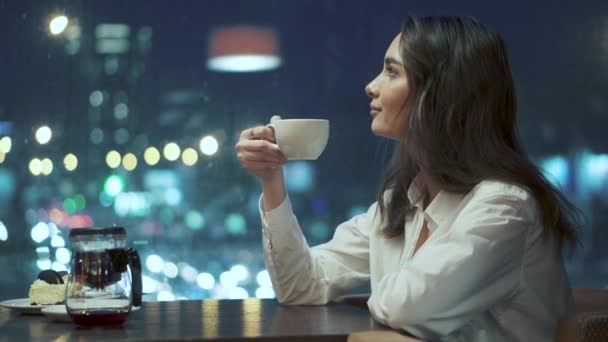 女孩喝茶, 并钦佩夜城的意见坐在一个舒适的咖啡馆深夜 — 图库视频影像