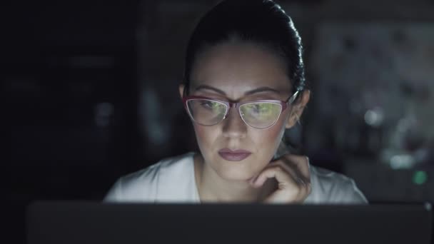 Η γυναίκα με τα γυαλιά δουλεύει σε ένα λάπτοπ στο σκοτάδι. πορτρέτο ενός κοριτσιού που περνά χρόνο στον υπολογιστή αργά το βράδυ. — Αρχείο Βίντεο