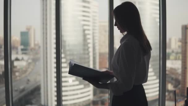 그녀의 손에 대 한 폴더와 매력적인 여자의 실루엣. 창문 가까이 사무실에서 서 있는 젊은 비즈니스 우먼 — 비디오