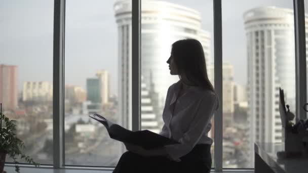 Silhouette di una donna che lavora in ufficio. ragazza si siede comodamente vicino alla finestra e lavora con i documenti — Video Stock