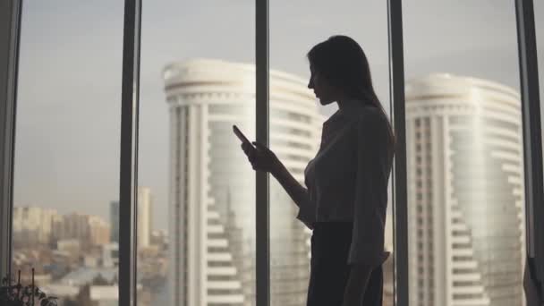 Silueta de una mujer joven sobre el fondo de una gran ventana y rascacielos. chica utiliza un teléfono inteligente . — Vídeo de stock