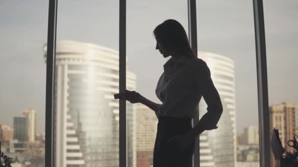 Silhouet van een jonge vrouw tegen de achtergrond van een groot raam en wolkenkrabber. het meisje is chatten op de telefoon. — Stockvideo