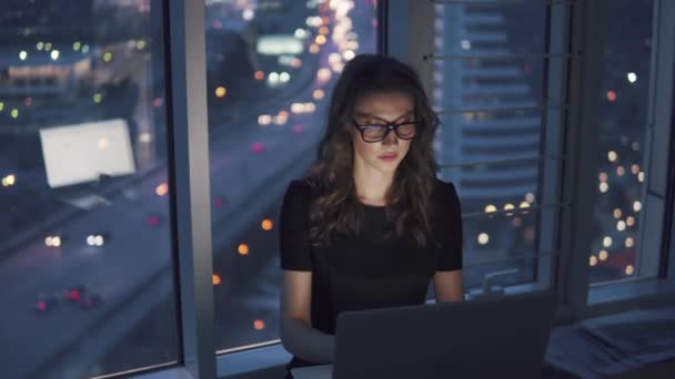 少女は、市内の夜のライトの背景に夜遅くにオフィスで動作します。メガネでモニターの反射を持つ若いビジネス女性 — ストック動画