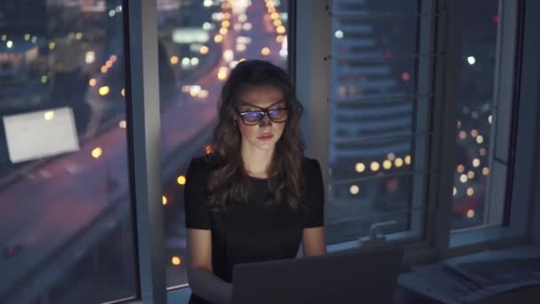 Praca późno w nocy w biurze. Młoda kobieta w garniturze i okulary działa na laptopie na tle miasta świateł w nocy. — Wideo stockowe