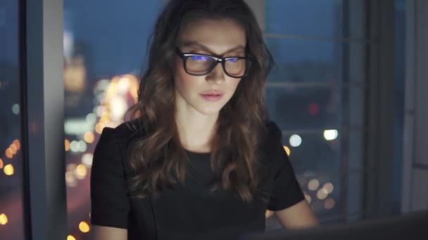 Працювати пізно ввечері в офісі. молода жінка в діловому костюмі і окулярах працює на ноутбуці на фоні нічних міських вогнів . — стокове відео