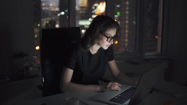 Retrato de una joven que trabaja hasta tarde en la oficina. mujer de negocios en gafas trabaja en un ordenador portátil en el fondo de la ciudad de la noche — Vídeo de stock