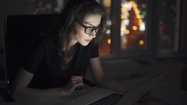 Retrato de una joven atractiva mujer de negocios que trabaja en una computadora portátil tarde en la noche en la oficina. chica en un traje de negocios y gafas en el fondo de las luces de la ciudad de la noche — Vídeos de Stock