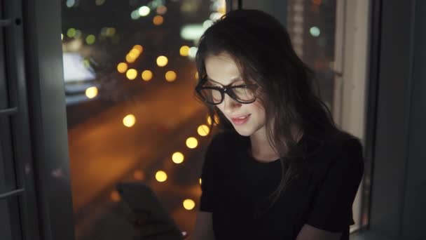 Retrato de una niña a la luz de la pantalla del teléfono inteligente. chica utiliza un teléfono móvil contra el fondo de la ciudad de la noche — Vídeo de stock