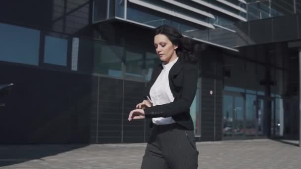 Femme d'affaires passe devant un centre d'affaires moderne. fille en costume d'affaires regarde l'horloge et se dépêche . — Video