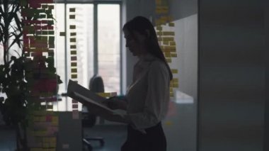 Elinde belgeler için bir klasör ile modern bir ofiste yürüyen bir kadının silüeti.