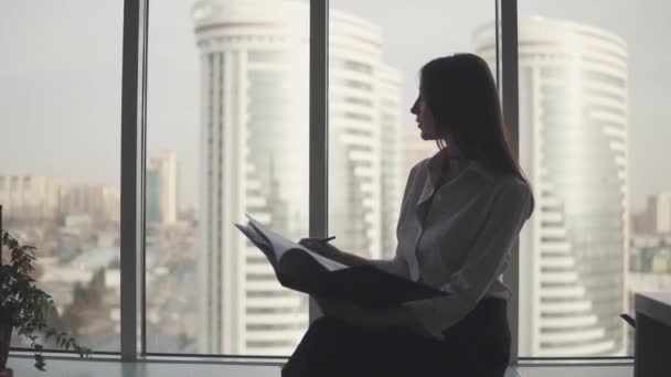 一个在办公室工作的女人的剪影。女孩舒舒服服地坐在靠窗的地方, 用文件工作 — 图库视频影像