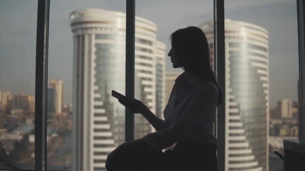 Силуэт девушки с телефоном в руках на фоне современных зданий. бизнес-леди пользуется смартфоном в офисе . — стоковое видео