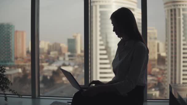 Young woman working on a laptop comfortably sitting near a large window. — Stock Video