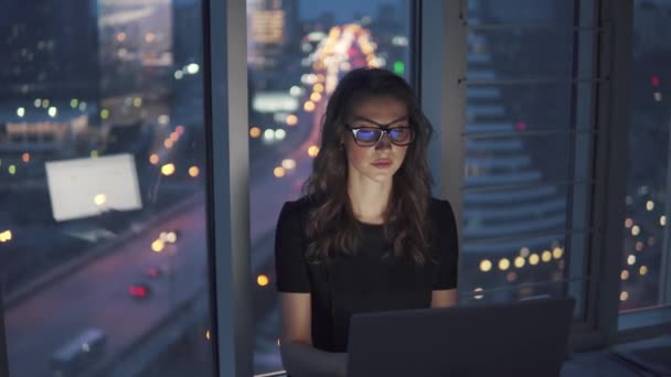 Gece geç saatte ofisinde çalışıyorum. genç kadın takım elbise ve gözlük gece şehir ışıkları arka plan üzerinde bir dizüstü bilgisayarda çalışıyor. — Stok video