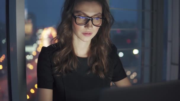 Zbliżenie portret kobiety biznesu na tle nocnego miasta. Dziewczyna pracuje późno w biurze. odbicie monitora w okularach — Wideo stockowe