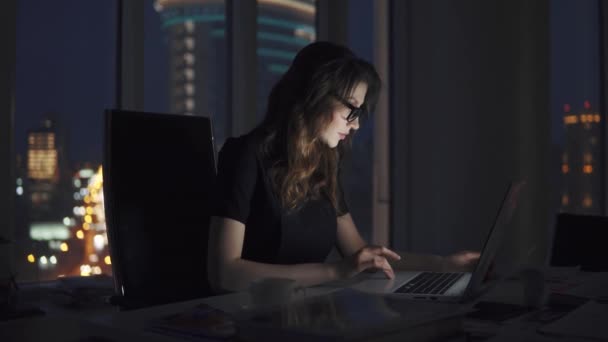 Una joven empresaria que trabaja hasta tarde en la oficina. retrato de una chica con gafas en el fondo de la ciudad nocturna — Vídeo de stock